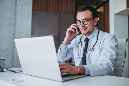 Young handsome doctor working with lapton. Talking on mobile phone and smiling.の写真素材