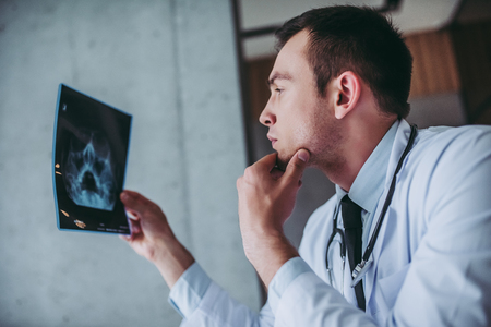 Young handsome doctor working. Serious doctor with stethoscope is looking at patient's x-ray image.の写真素材