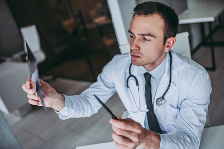 Young handsome doctor working. Serious doctor with stethoscope is looking at patient's x-ray image.の写真素材