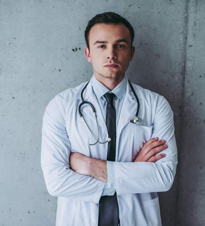 Portrait of young handsome doctor standing on grey background with stethoscope.の写真素材