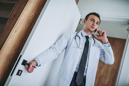 Young handsome doctor with stethoscope working. Opening the door and talking on mobile phone.の写真素材