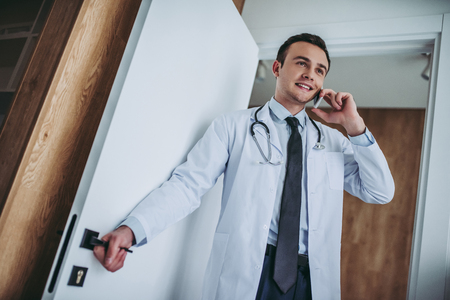 Young handsome doctor with stethoscope working. Opening the door, talking on mobile phone and smiling.の写真素材