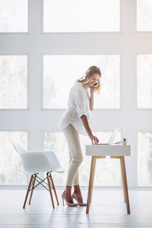 Full-length image of attractive pregnant woman is working with laptop in light modern office and talking on mobile phone. Last months of pregnancy.の写真素材