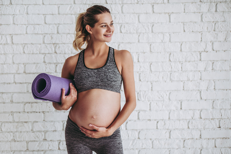 Beautiful pregnant woman workout. Doing fitness on last months of pregnancy. Yoga. Standing on white brick background with sports mat.の写真素材