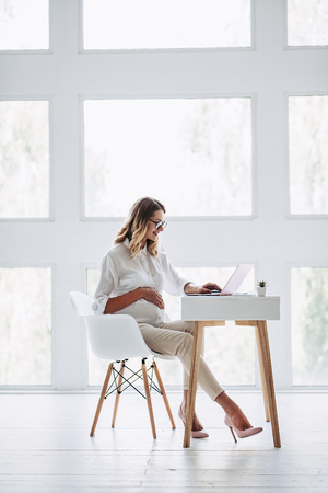 Full-length image of attractive pregnant woman is working with laptop in light modern office. Last months of pregnancy.の写真素材