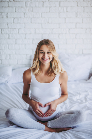 Attractive pregnant woman is sitting in bed and making the heart sign on belly. Last months of pregnancy.の写真素材