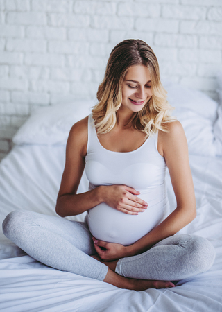 Attractive pregnant woman is sitting in bed and holding her belly. Last months of pregnancy.の写真素材