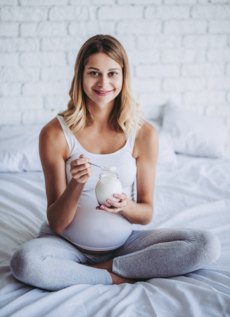 Attractive pregnant woman is sitting in bed and eating yogurt. Healthy food concept. Last months of pregnancy.の写真素材