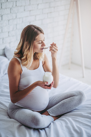 Attractive pregnant woman is sitting in bed and eating yogurt. Healthy food concept. Last months of pregnancy.の写真素材