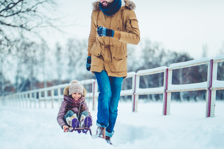 Handsome young dad and his little cute daughter are having fun outdoor in winter. Enjoying spending time together while riding on a sled. Family concept.の写真素材