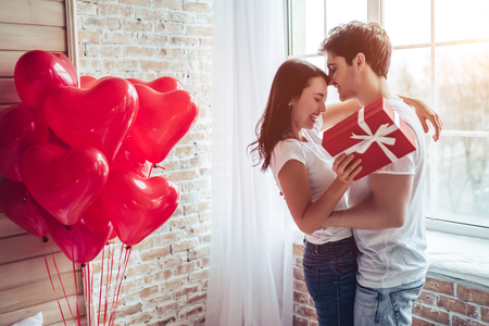 Beautiful young couple at home. Hugging, kissing and enjoying spending time together while celebrating Saint Valentine's Day with gift box in hand and air balloons in shape of heart on the background.