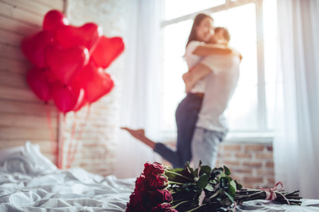 Beautiful young couple at home. Hugging, kissing and enjoying spending time together while celebrating Saint Valentine's Day with red roses on bed and air balloons in shape of heart on the background.