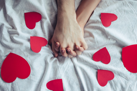 Cropped image of young couple is lying on bed with red hearts nearby. Holding hands. Saint Valentines Day.の写真素材