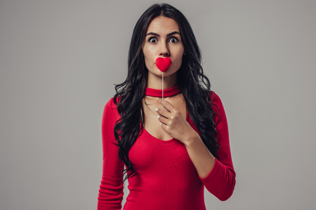 Portrait of attractive young woman is posing on grey background with red heart in hands.の写真素材