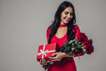 Portrait of attractive young woman is standing on grey background with gift box and red roses in hands.の写真素材
