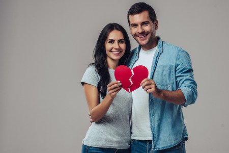 Beautiful romantic couple is standing on grey background with cardboard red heart in hands. Happy Saint Valentine's Day!の写真素材