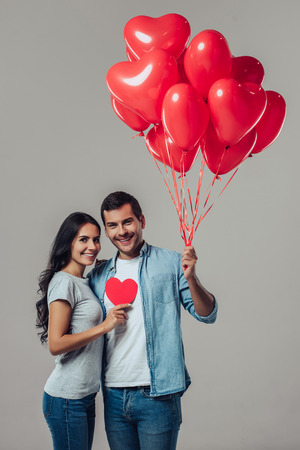 Beautiful romantic couple isolated on grey background. Attractive young woman and handsome man are hugging and smiling with air balloons in shape of heart in hands. Happy Saint Valentine's Day!の写真素材