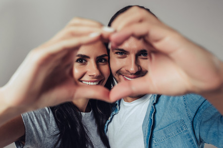 Beautiful romantic couple isolated on grey background. Attractive young woman and handsome man are smiling and looking at camera while making a heart sign with their hands.の写真素材