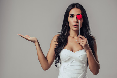 Portrait of attractive young woman is standing on grey background with red heart in hand.の写真素材