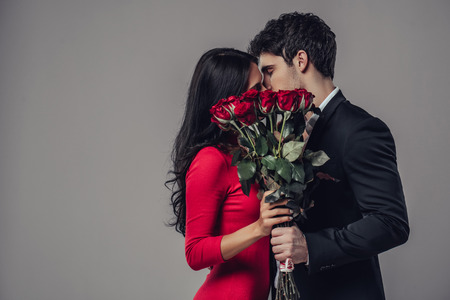 Beautiful romantic couple isolated on grey background. Attractive young woman in dress holding red roses and handsome man in suit are kissing. Happy Saint Valentine's Day!の写真素材