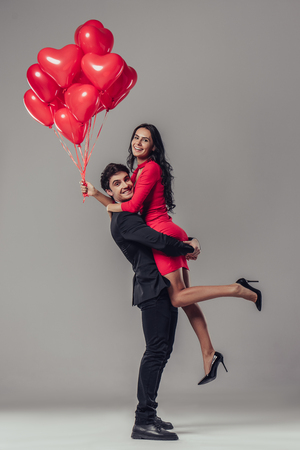 Beautiful romantic couple isolated on grey background. Attractive young woman in red dress and handsome man in suit are hugging with air balloons in hands. Happy Saint Valentine's Day!の写真素材