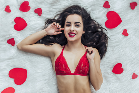 Top view of young sexy woman is lying on bed in red lingerie with red heart in hand and smiling. Happy Saint Valentine's Day!の写真素材