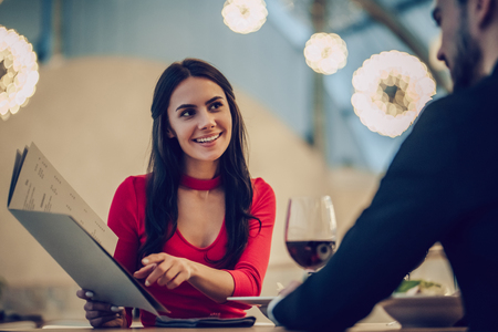 Beautiful loving couple is spending time together in modern restaurant. Attractive young woman in dress and handsome man in suit are having romantic dinner. Celebrating Saint Valentine's Day.の写真素材