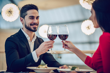 Beautiful loving couple is spending time together in modern restaurant. Attractive young woman in dress and handsome man in suit are having romantic dinner. Celebrating Saint Valentine's Day.の写真素材