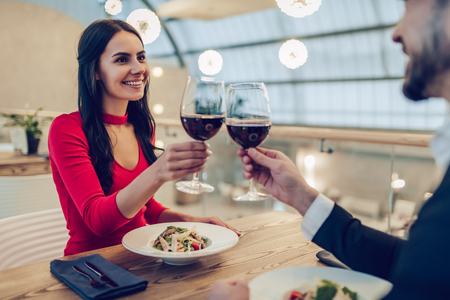 Beautiful loving couple is spending time together in modern restaurant. Attractive young woman in dress and handsome man in suit are having romantic dinner. Celebrating Saint Valentine's Day.の写真素材