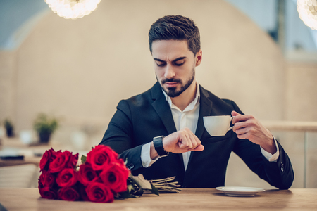 Handsome businessman is sitting in modern restaurant with bouquet of red roses and checking the time while waiting for his beloved.の写真素材