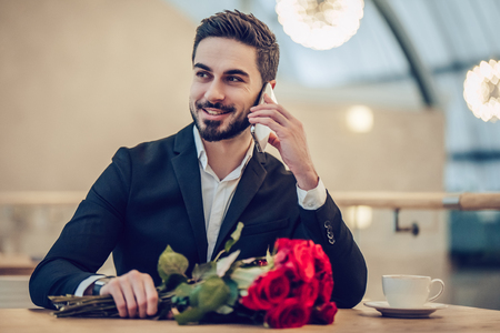 Handsome businessman is sitting in modern restaurant with bouquet of red roses and talking on mobile phone while waiting for his beloved.の写真素材