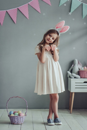 Little cute girl with bunny ears is standing with basket full of colorful Easter eggs while preparing for Easter celebration.の写真素材