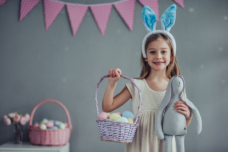 Little cute girl with bunny ears is standing with basket full of colorful Easter eggs while preparing for Easter celebration.の写真素材