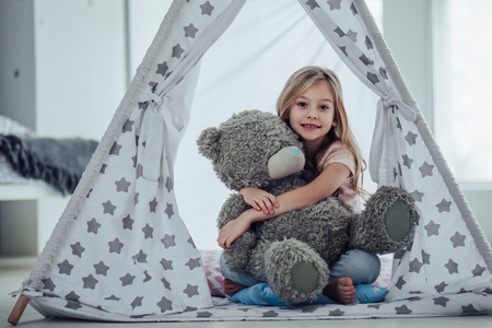 Little cute girl is sitting on the floor at home, hugging teddy bear, smiling and looking at camera.