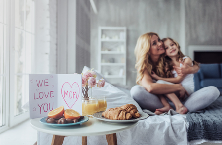 Attractive young woman with little cute girl are spending time together at home while sitting on bed. Happy family concept. Breakfast in bed on Mother's day.の写真素材