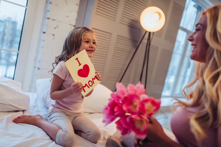 I love you, mom! Attractive young woman with little cute girl are sitting on bed and spending time together at home. Mom is receiving presents from daughter on Mother's Day. Happy family concept.の写真素材