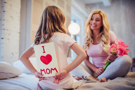I love you, mom! Attractive young woman with little cute girl are sitting on bed and spending time together at home. Mom is receiving presents from daughter on Mother's Day. Happy family concept.の写真素材