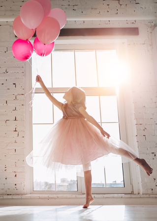 Cute little girl in beautiful dress is dancing at light sunny room with pink air balloons in hand.の写真素材