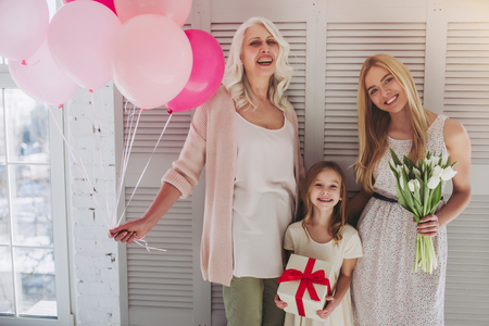 Little cute girl, her attractive young mother and charming grandmother are standing with air balloons and flowers in light room. Women's generation. International Women's Day. Happy Mother's Day.の写真素材