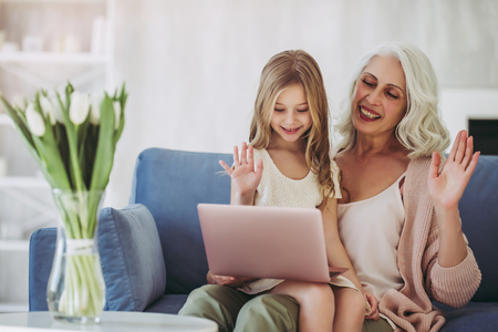 Little cute girl and her grandmother are spending time together at home. Sitting on sofa with laptop and waving handsの写真素材