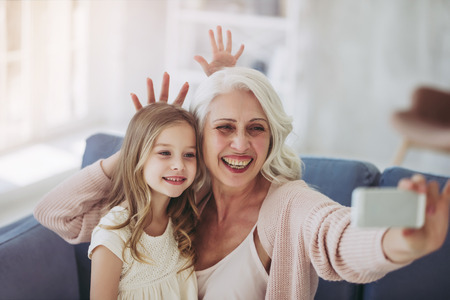 Little cute girl and her grandmother are spending time together at home. Having fun while sitting on sofa and making selfie on a smart phoneの写真素材