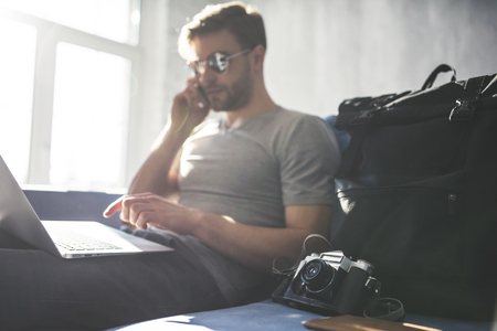 Handsome young man is sitting at home with smart phone and laptop. Preparing for travelingの写真素材