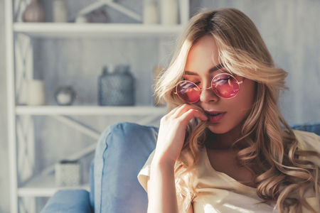 Portrait of attractive young woman is sitting on sofa at home in pink sunglassesの写真素材