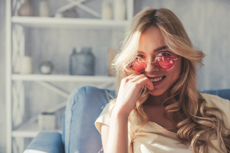 Portrait of attractive young woman is sitting on sofa at home in pink sunglassesの写真素材