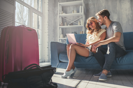 Young romantic couple is preparing for travel at home. Sitting on sofa with laptop, passports and tickets in hands while suitcases are standing nearbyの写真素材