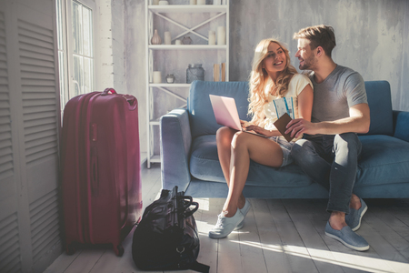 Young romantic couple is preparing for travel at home. Sitting on sofa with laptop, passports and tickets in hands while suitcases are standing nearbyの写真素材