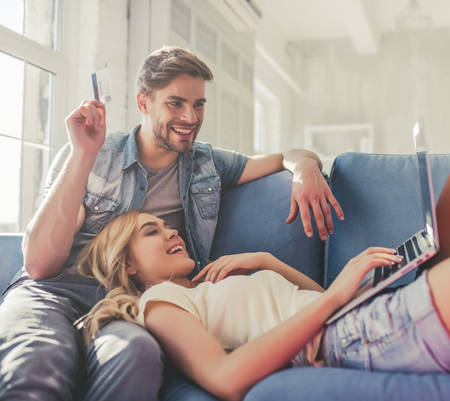 Handsome man is sitting on sofa with credit card in hand while attractive young woman is lying on his knees with laptop. Online shopping.の写真素材