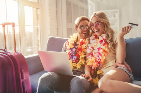 Young romantic couple wearing Hawaii accessories and sunglasses is preparing for travel at home. Sitting on sofa with laptop and credit card in hands while suitcases are standing nearbyの写真素材