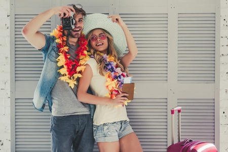 Young romantic couple is preparing for traveling. Standing on grey background in Hawaii accessories, sunglasses and hat with passport and ticket in hand. Suitcases are nearby.の写真素材