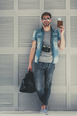Handsome young man is preparing for traveling. Standing on grey background with suitcase and retro camera in sunglasses with passport and ticket in hand.の写真素材